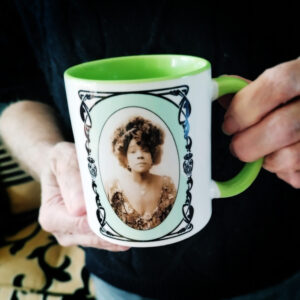 A ceramic mug featuring a sepia-tone portrait of Aida Overton Walker, framed by an elegant Art Deco border on a soft green background. The mug has a matching green handle and interior, and is being held by a person in a dark sweater.