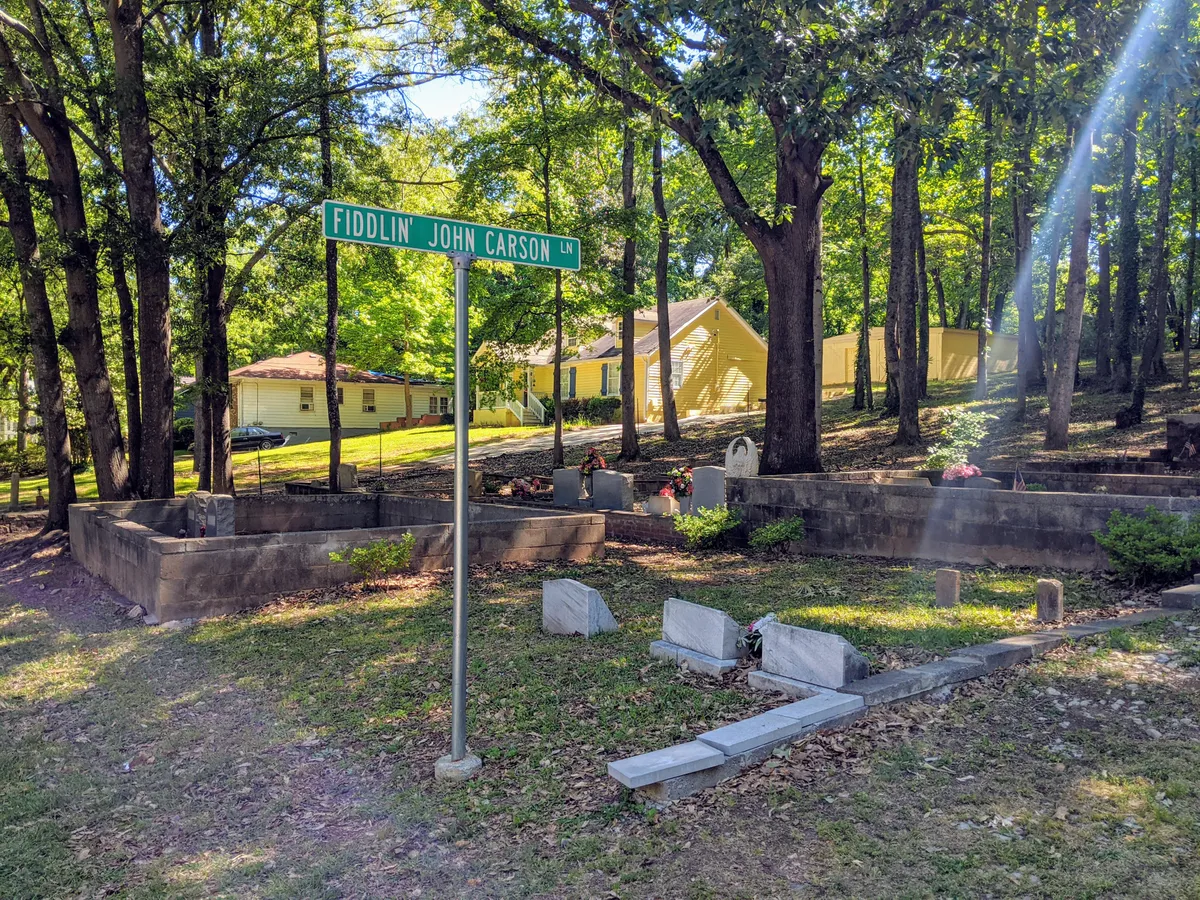 Gravesite of Fiddlin’ John Carson in Atlanta, Georgia, with a street sign reading “Fiddlin’ John Carson Ln” amidst trees and gravestones.